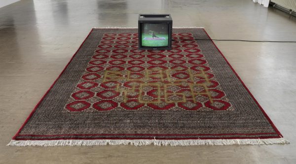 A small tv on a Persian-style rug displaying a person doing yoga on a similar rug in a park
