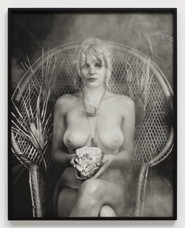 A naked, feminine person holding a rock sits in a chair.
