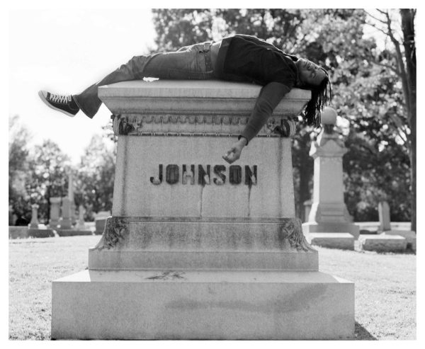 Black and white photo of Rashid Johnson lying on top of a tombstone labeled Johnson