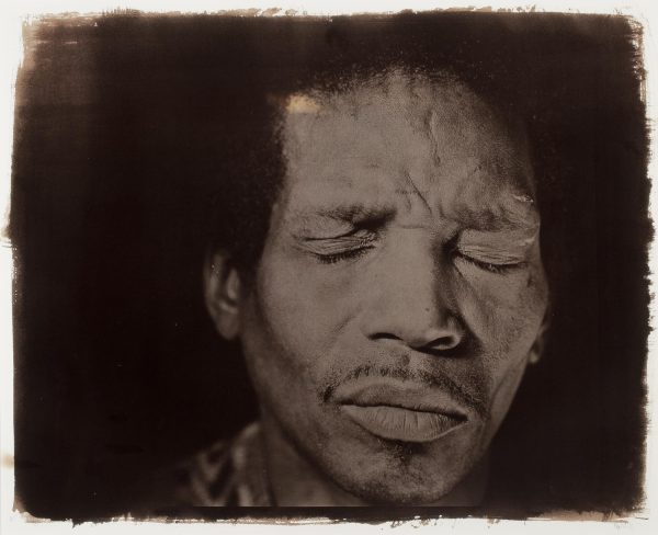 A brown-toned photograph featuring a close-up of a male-presenting person with their eyes closed