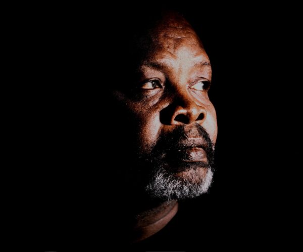 A headshot captures a dark-skinned person with a black mustache and short white beard looking off towards their left, with a black background.