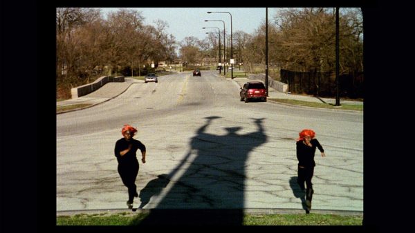 An elongated shadow of a person holding a broom as they raise both hands is cast between two people dressed identically in a red hat and black shirt and pants on an empty street