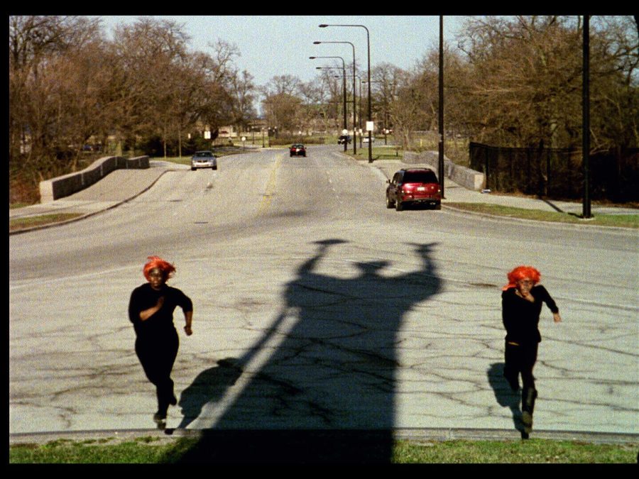 An elongated shadow of a person holding a broom as they raise both hands is cast between two people dressed identically in a red hat and black shirt and pants on an empty street