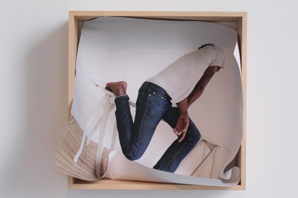 A photo of a person with medium-brown skin bending their legs and twisting their upper body is shoved into a square wooden box