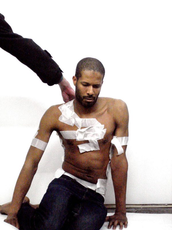 A dark-skinned person with white tape covering small portions of their chest and arms sits on the floor as another person's arm reaches for something on their back