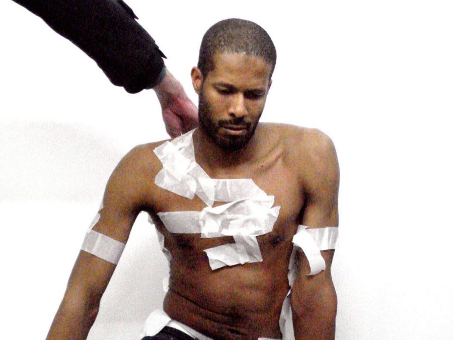A dark-skinned person with white tape covering small portions of their chest and arms sits on the floor as another person's arm reaches for something on their back