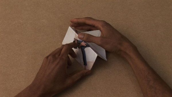 A pair of medium-dark skinned hands fold a picture of a person into a three dimensional shape