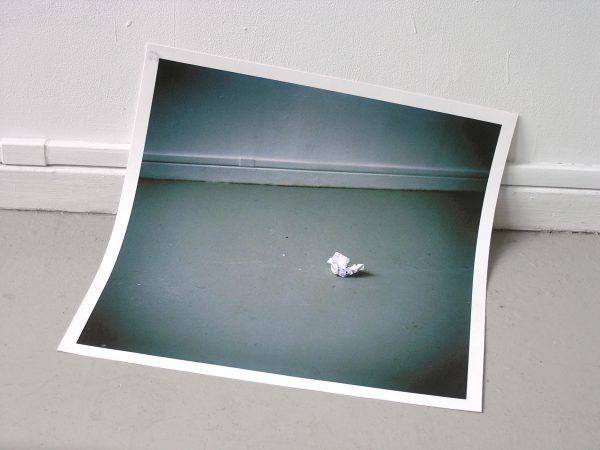 A black and white picture of a crunched up wad of paper near a baseboard is folded to line up with a tan baseboard and floor