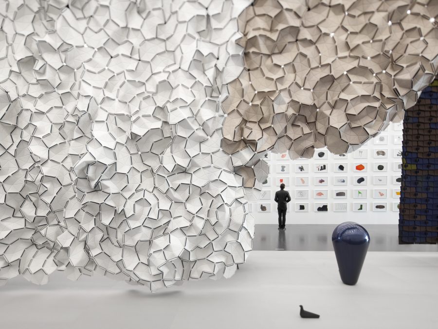 A giant cloud-like installation of white and tan bean-shaped objects divide a gallery into two spaces. A person looks at a wall of objects in the distance.