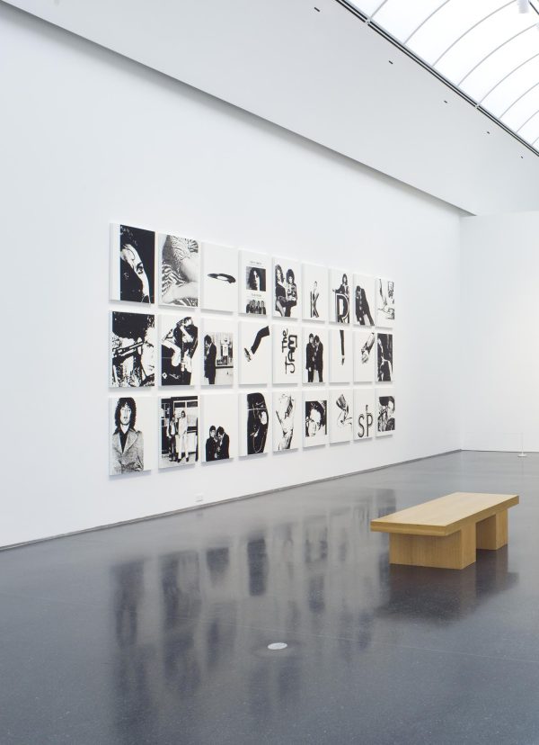 A wooden bench in front of a series of black and white paintings displayed in three rows of nine in a vaulted gallery space
