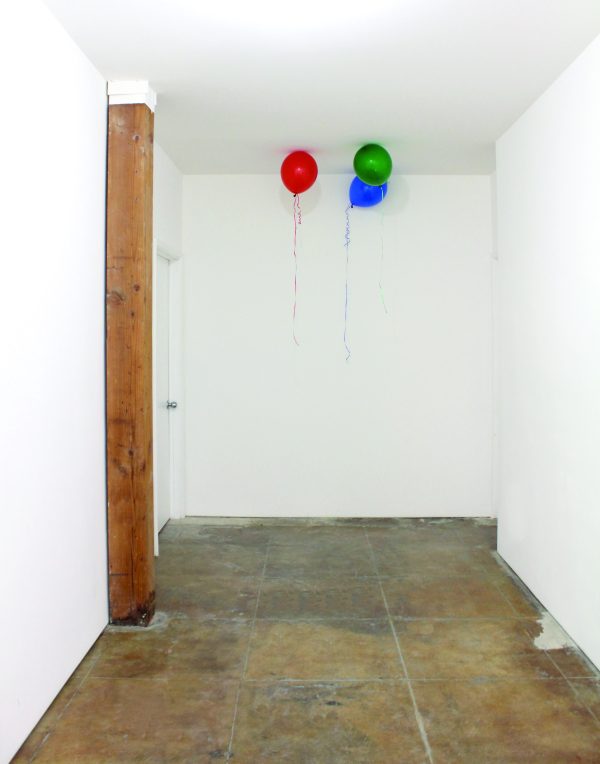 Three colorful helium balloons rest against the ceiling of a white hallway with a brown stone floor and vertical wood beam against its wall, frame left.