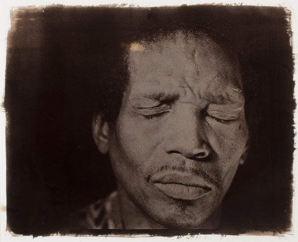 A brown-toned photograph featuring a close-up of a male-presenting person with their eyes closed