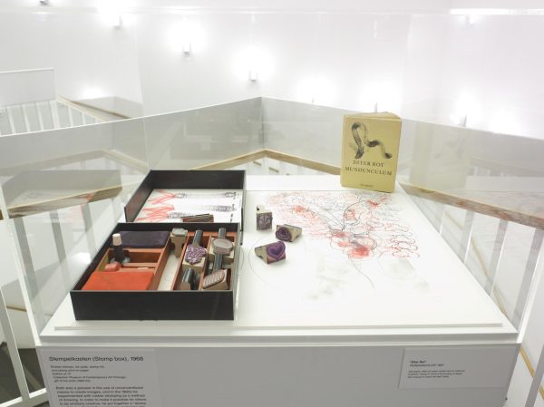 Display case near a stairwell with white banisters featuring a stamp kit with a few stamps out and examples of their imprints on paper