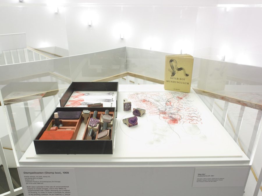 Display case near a stairwell with white banisters featuring a stamp kit with a few stamps out and examples of their imprints on paper