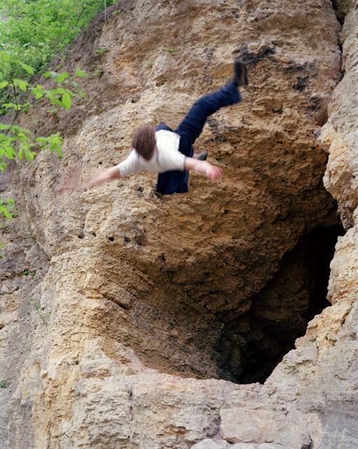 A pale-skinned person is mid-fall in front of a gaping stone cave.