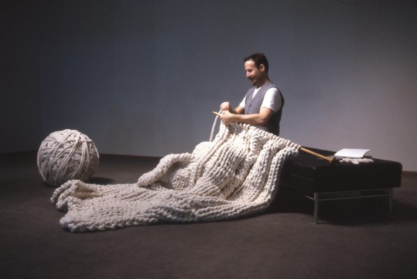 A person sits on a bench knitting a large cream blanket from a giant ball of yarn