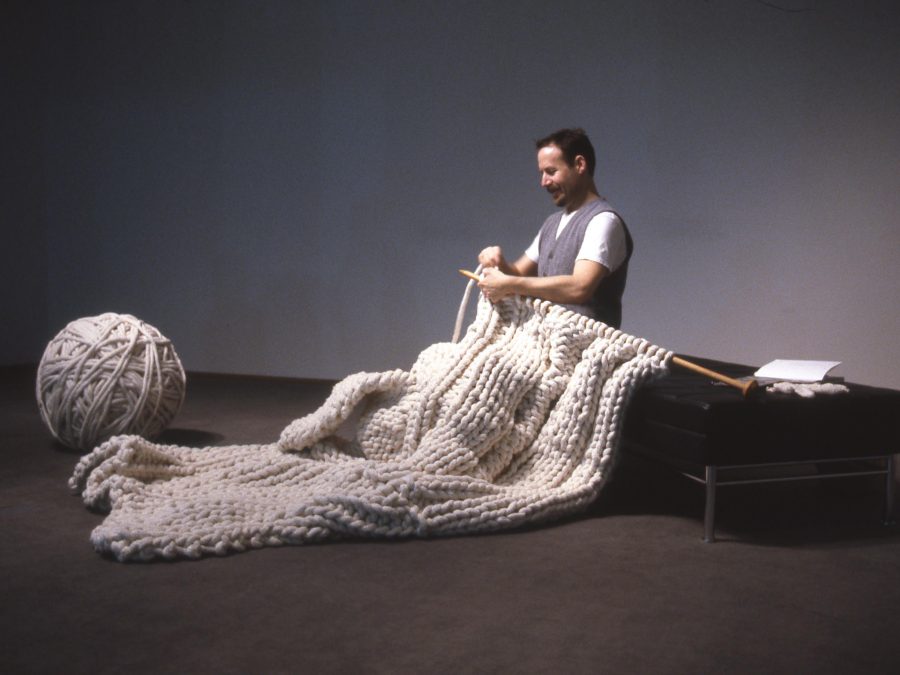 A person sits on a bench knitting a large cream blanket from a giant ball of yarn