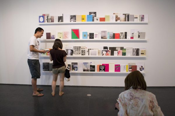 Two pale-skinned people are standing in front of a wall of shelves with books. Another person with their back to you sits in the foreground.