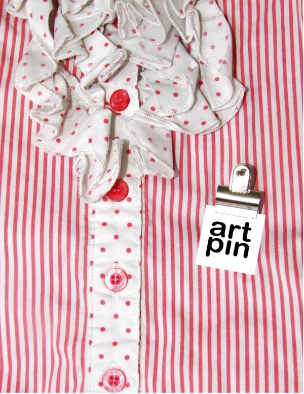 A red and white striped blouse with a red polka dot ruffled collar and button seam with a small clip on the left side displaying large black text on a white square that reads art pin