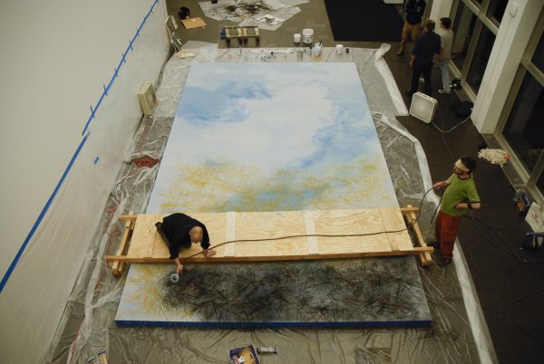 A person kneeling on a board spanning a giant canvas spray paints a small section black