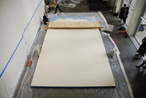 From a high viewpoint you see a couple of people walking around a giant canvas resting on a sheet of plastic. The furthest section of the canvas has been painted blue