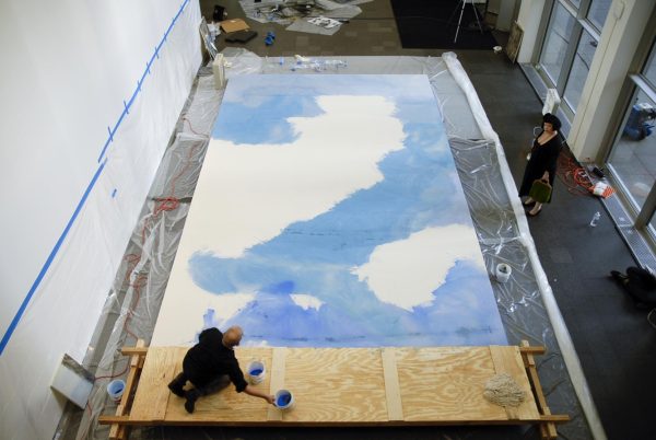 A person sits on a board spanning a giant canvas; the majority of the canvas features a blue section that resembles a backwards s