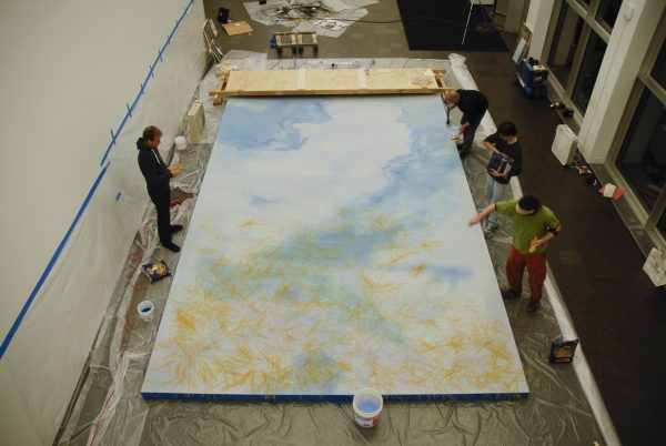 A group of people scatter thin yellow objects on a giant canvas partially painted blue in a way that resembles clouds in the sky