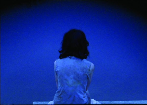 A deep blue–toned photo shows a person with shoulder-length black hair sitting with their back to the viewer.