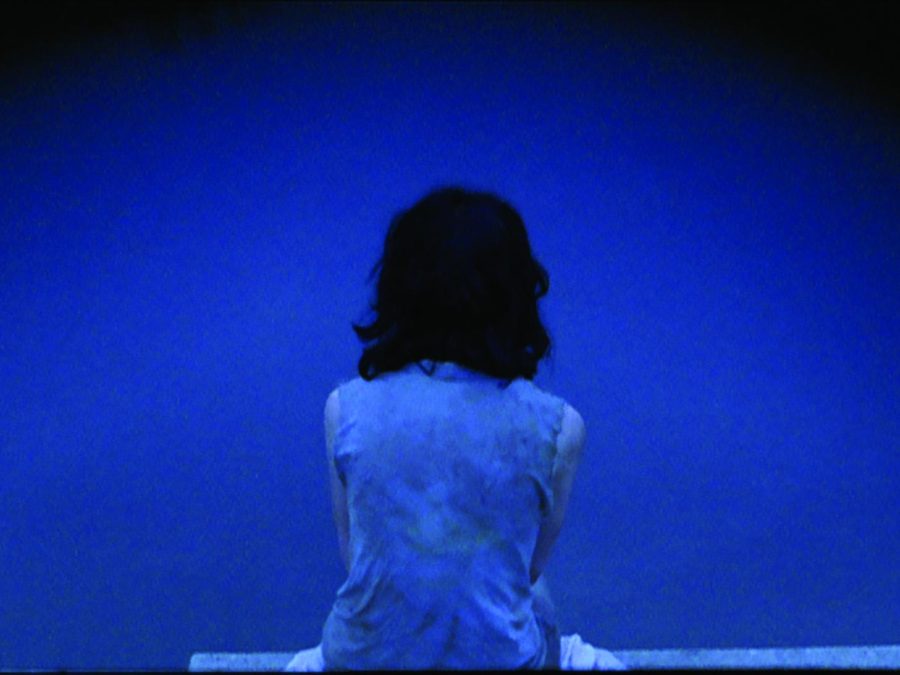 A deep blue–toned photo shows a person with shoulder-length black hair sitting with their back to the viewer.