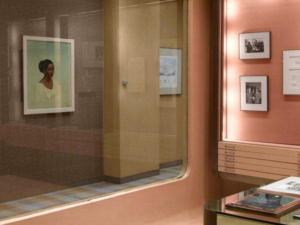 A view into a hallway with works hanging on the wall from a window in a peach room with drawers and a glass desk just visible from the bottom right