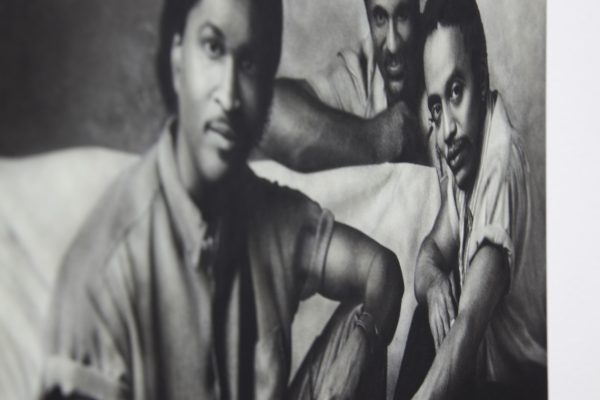 Distorted black and white image of three male-presenting people with medium-dark skin sitting near a piece of furniture covered in white fabric