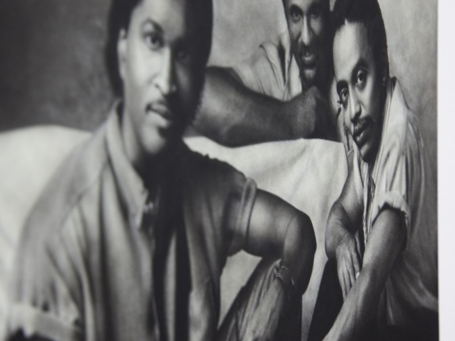 Distorted black and white image of three male-presenting people with medium-dark skin sitting near a piece of furniture covered in white fabric