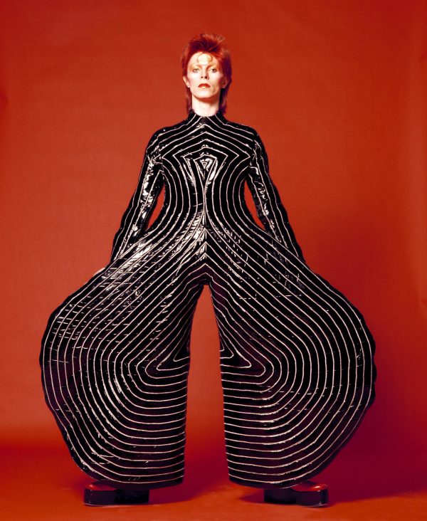 A photograph of David Bowie with pale skin and bright red hair wearing a wide-leg black jumpsuit with white stripes in front of a red background.