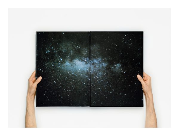 Two arms hold a book open to a page displaying an image of a galaxy on a white table