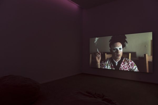 Still of someone in a robe smoking. They have white makeup around their eyes.