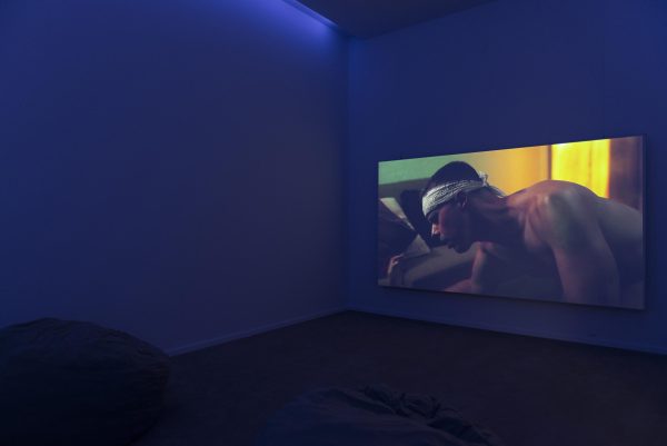 Still of a topless masculine-looking person with a bandana tied around his head seemingly crawling on the floor.
