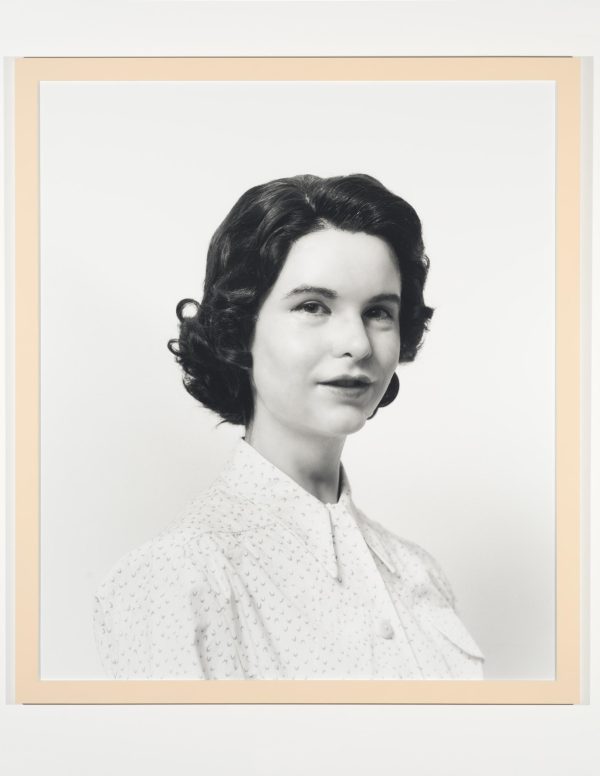 A black-and-white photograph portrait shows a pale-skinned person with short dark hair wearing a white blouse in front of a white background.