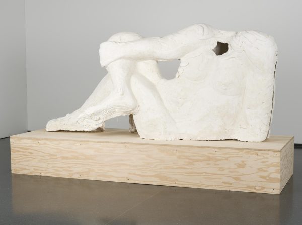 A white plaster case of a human figure with no head in a relaxed reclined pose on a wooden platform