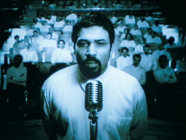 A teal-tinted image of a man with a goatee standing in front of a microphone with a crowd behind him in tiered rows