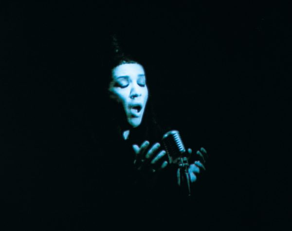 A dark vignette surrounds a person with female features cast in a blue light and passionately singing into a microphone