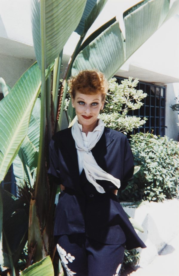 A female presenting person dressed like Lucille Ball near a large fern