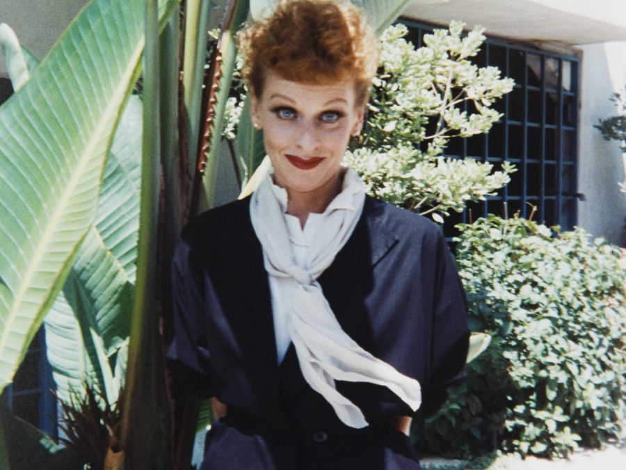 A female presenting person dressed like Lucille Ball near a large fern