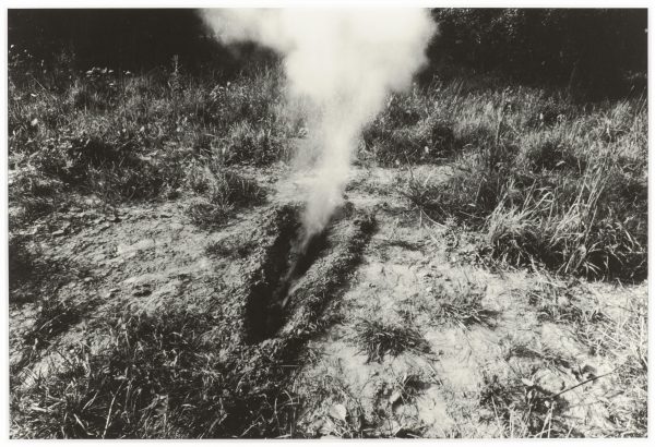 A black-and-white photograph depicts smoke billowing from a body-sized trench in a clearing with weeds and trees on the horizon.