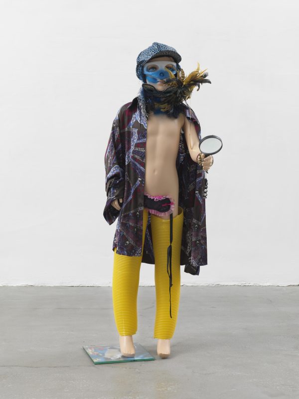 A male mannequin with light skin holding a magnifying glass and dressed in yellow pants, an open coat, a boa, goggles, and a leather cap
