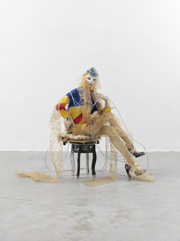 A light-skinned mannequin sitting on a small stool with its legs crossed. It wears a white mask and a yellow, red, and blue rain coat. It is wrapped in cream lace fabric that spills to the floor.
