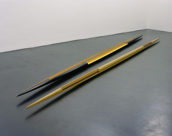 Two partially painted wooden spears with points on both ends on a grey floor
