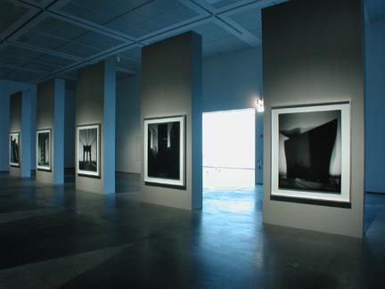In a darkened room a row of grey, floor-to-ceiling wall dividers display one photograph each