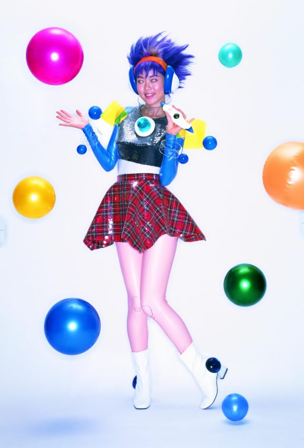 A futuristic person wearing a brightly colored crop top and a skirt poses in between thirteen colorful spheres.