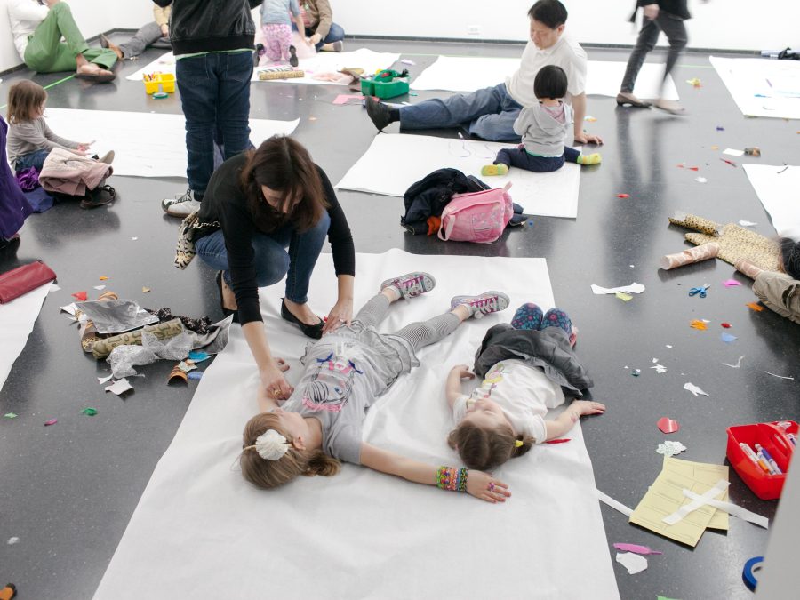 Parents and children interacting together in a white-walled gallery, with children lying down on large sheets of white craft paper while their parents trace the outline of their bodies.