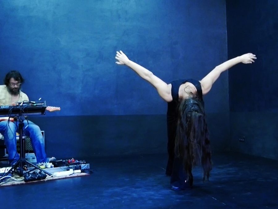 Surrounded by deep blue walls and floor, a person with long hair stands with arms out, bending very far forward while to their right another person plays guitar.